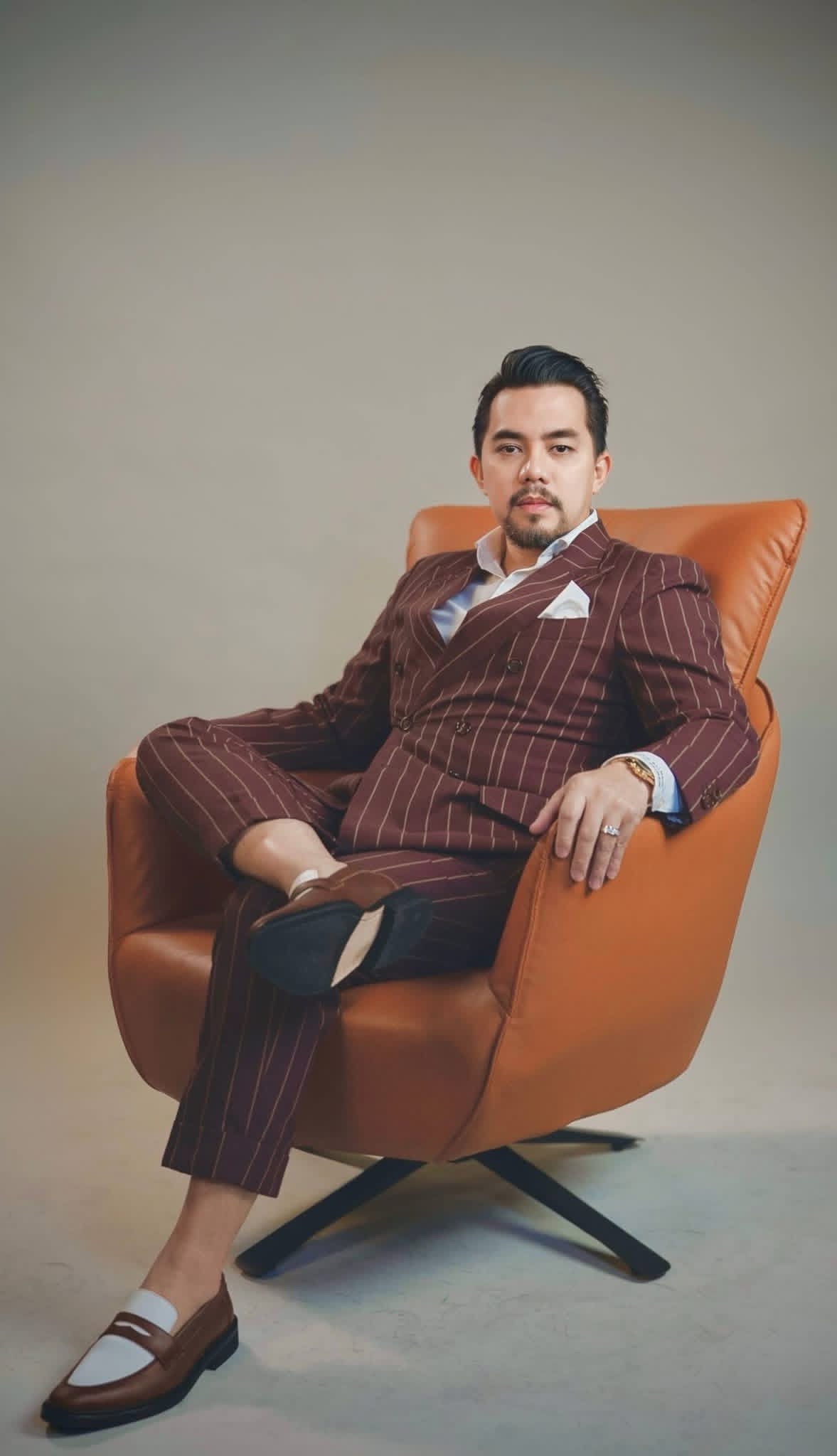 Editorial portrait of Reymond delos Reyes seated in a maroon pinstripe suit on an orange leather chair
